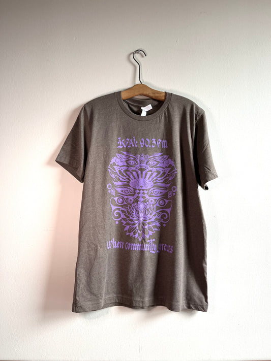 Heather brown short sleeve tee shirt with lilac purple decorative design on the front. The design is a mirror image down the center with two sets of eyes, lotus-style flower, and other accents. The text on the shirt says: KFAI 90.FM and Where Community Grows.