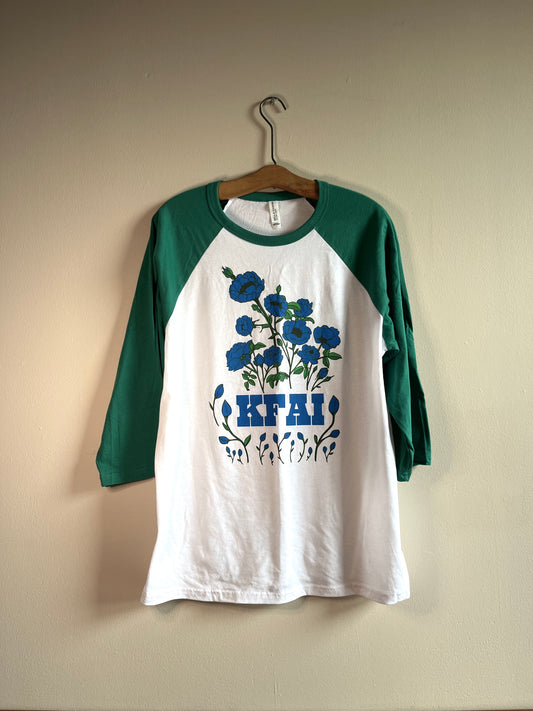 Baseball style 3/4 sleeve tee shirt. Kelly green sleeve and white shirt body. The graphic is a large stylized illustration of blue roses with block KFAI letters set in the bottom 1/4 of the design.