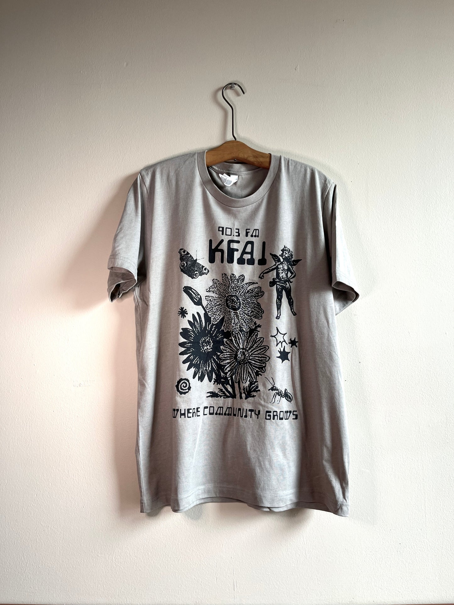 Beige shirt with black ink. The design is comprised of collage elements of flowers, angels, and insects. The text reads 90.3FM KFAI and Where Community Grows.